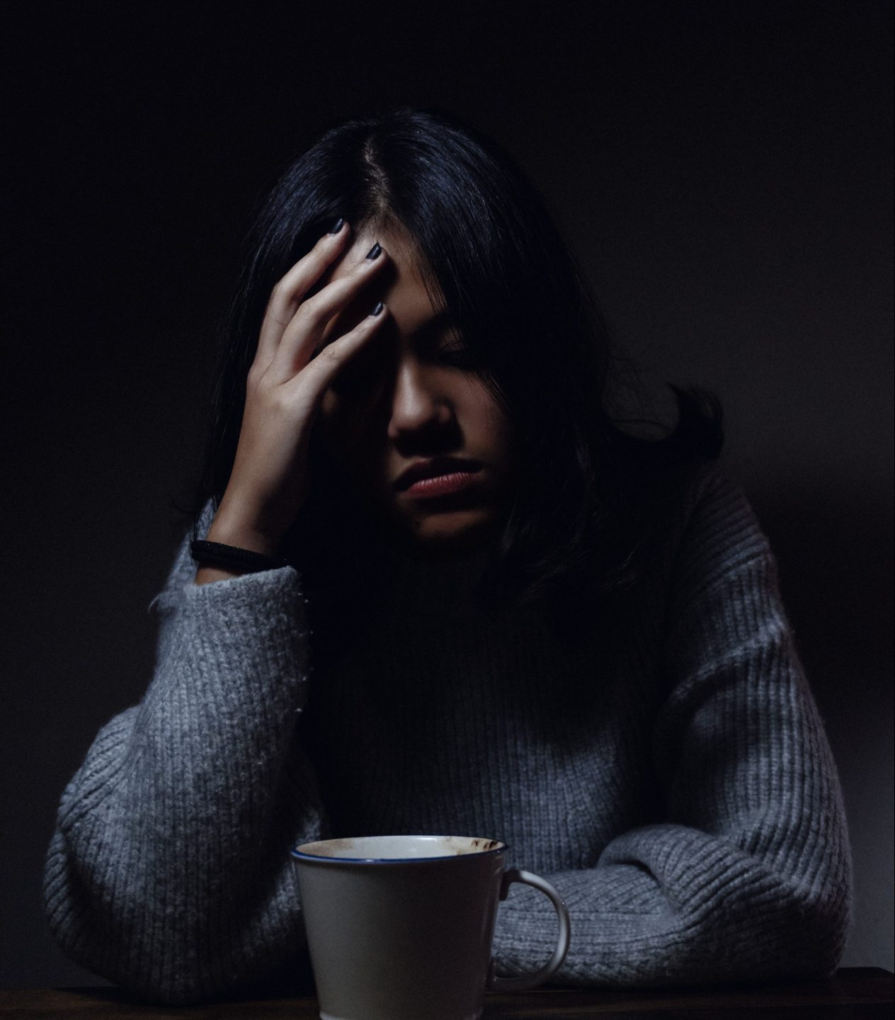 Visibly upset woman holding her head in her hands while sitting at a table with a mug